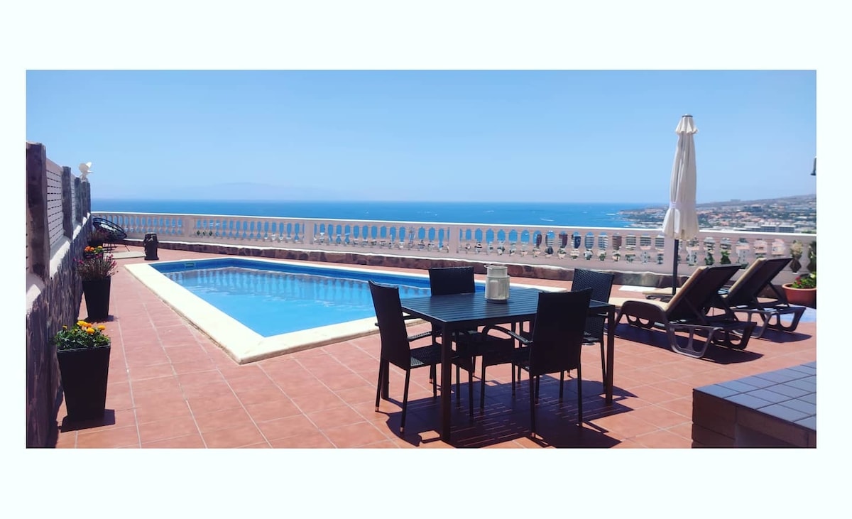 An outdoor terrace features a blue swimming pool surrounded by sun loungers and a dining table. Potted plants add greenery, while an umbrella provides shade. The expansive view showcases the ocean and coastline under a clear blue sky.
