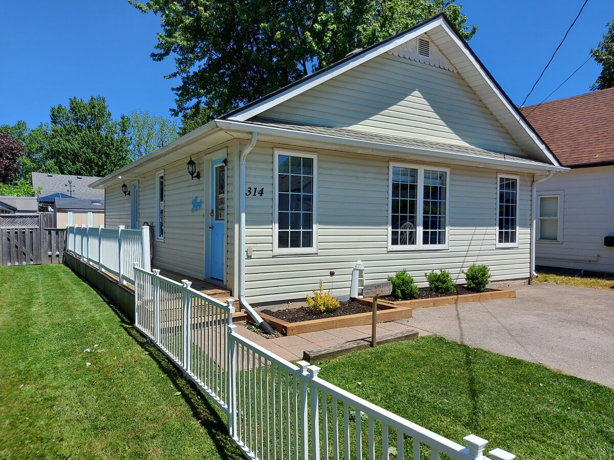 A charming exterior of a single-story cottage is displayed, featuring light-colored siding and multiple large windows. A white picket fence outlines the landscaped yard, which includes a green lawn and small shrubs framing the front entrance. The bright blue front door adds a welcoming touch.