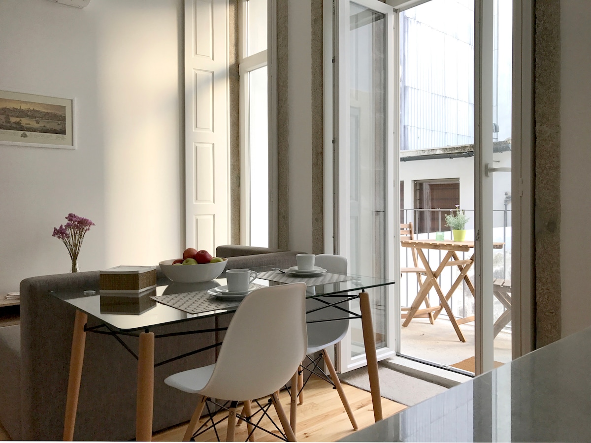 A dining area is showcased, featuring a glass-topped table paired with white chairs. A bowl of apples sits on the table, and sunlight filters through large glass doors leading to a balcony. A cozy sofa is visible in the background, enhancing the open space feeling.
