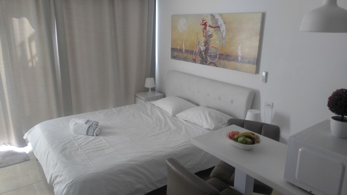 The room features a spacious bed with crisp white bedding, complemented by a decorative wall art piece. A small table holds a bowl of fruit, and a kitchenette is visible nearby. Soft, neutral curtains frame the window, allowing filtered light to enter.
