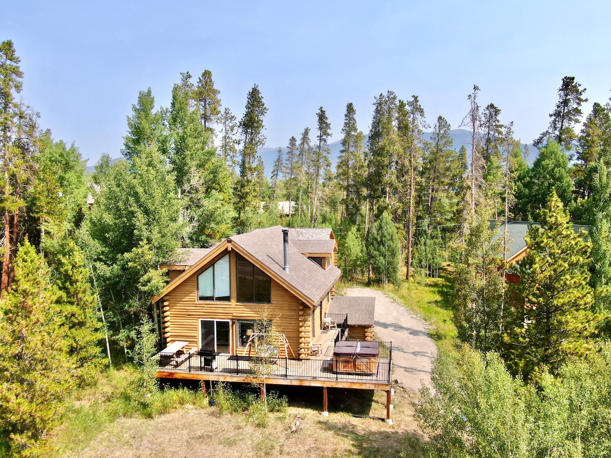 A spacious mountain retreat is nestled among tall trees, with its wooden exterior blending into the natural surroundings. The deck features a private hot tub, and large windows allow for a view of the mountains in the distance.