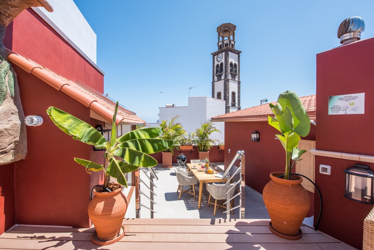 The private terrace is adorned with potted plants and contemporary dining furniture. A spacious table is flanked by comfortable chairs, inviting outdoor meals. In the background, a clock tower can be seen rising above the rooftops against a clear blue sky.