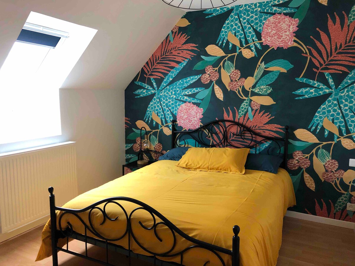 A vibrantly decorated bedroom features a floral mural on one wall, complemented by a spacious bed with a yellow duvet. Natural light enters through a slanted window, and a radiator is positioned beneath it, adding to the cozy ambiance of the room.