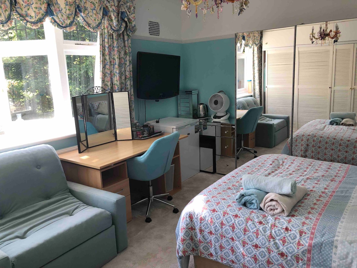 A well-lit room features a spacious bed with decorative bedding, complemented by a comfortable seating area and a writing desk. A mirror reflects the arrangement, while a flat screen TV is mounted on the wall. Large windows allow natural light to illuminate the space.