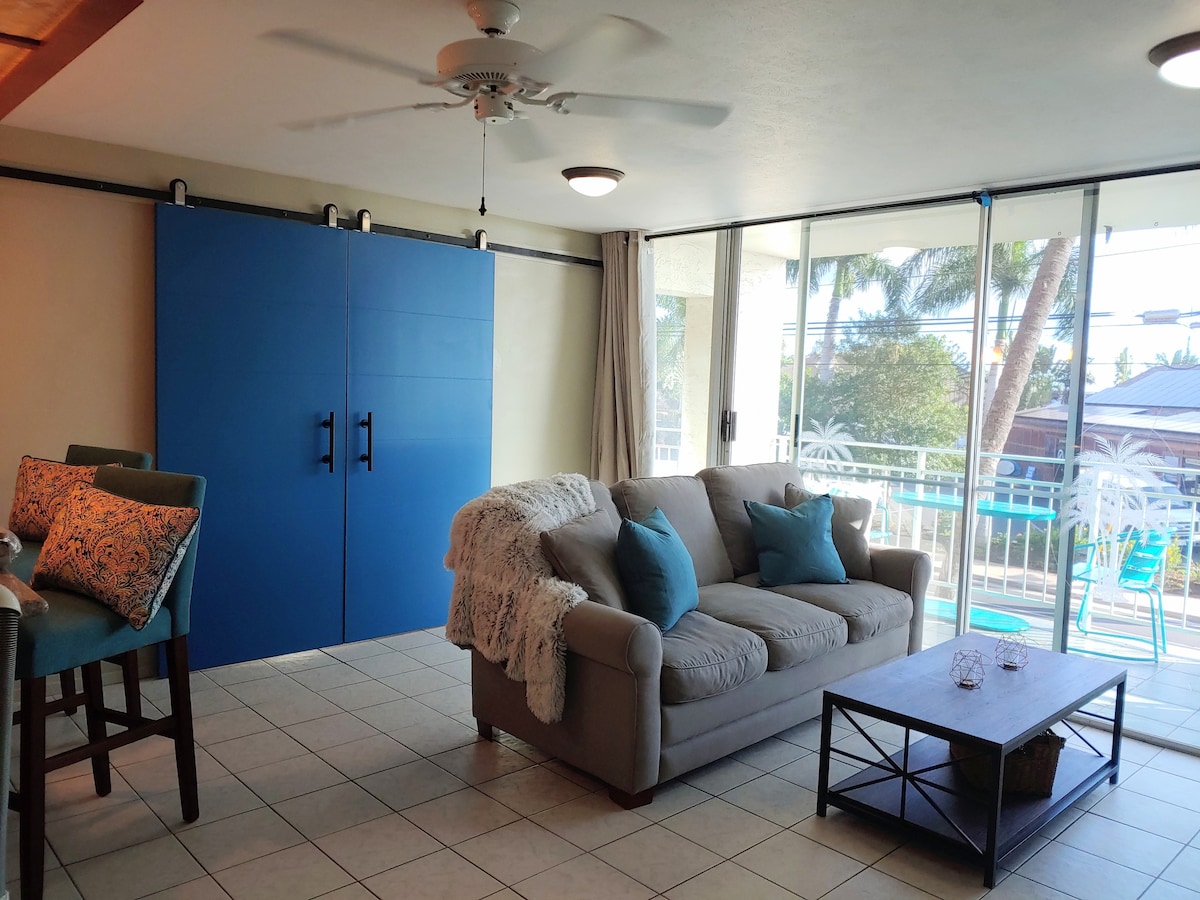 A spacious living area features a light gray sofa adorned with colorful throw pillows, positioned adjacent to a low coffee table. A large sliding glass door provides access to the balcony, with views of palm trees and a pool area visible beyond.