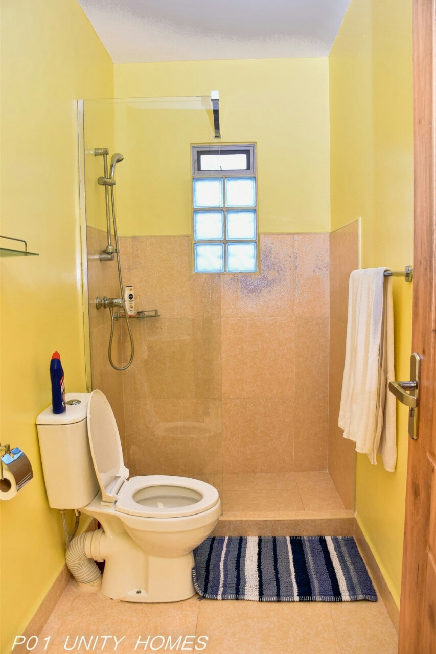 A bright bathroom features yellow walls and a glass shower enclosure. A modern toilet is positioned next to a small towel rack. A striped bath mat lies on the floor, with a window allowing natural light to illuminate the space.
