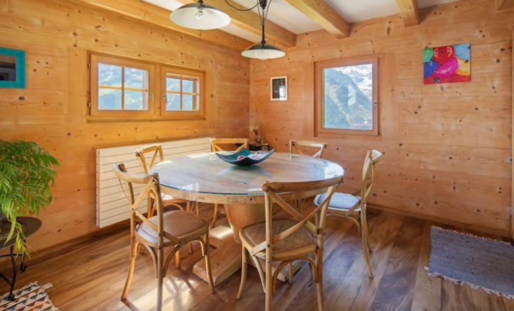 A wooden dining area features a round table surrounded by six chairs. Natural light enters through two windows, illuminating the rustic interior. Colorful artwork adorns the walls, adding a touch of character to the space.