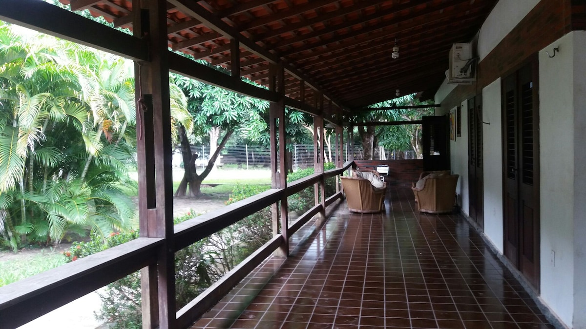 A covered porch features a rustic wooden ceiling and tiled flooring. Comfortable seating is provided with two armchairs arranged to face the lush greenery beyond. Views of the surrounding natural landscape are framed by wooden beams, creating a calm and inviting space for relaxation.