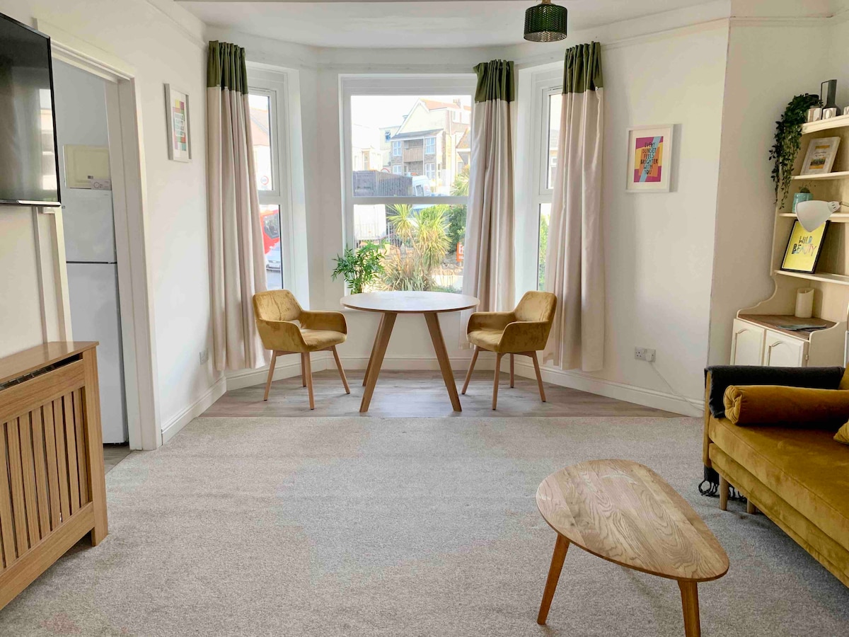 The front room features large bay windows allowing ample natural light. A circular dining table is surrounded by two upholstered chairs. A low wooden coffee table is positioned in front of a cozy sofa, with soft carpeting enhancing the space's warmth.