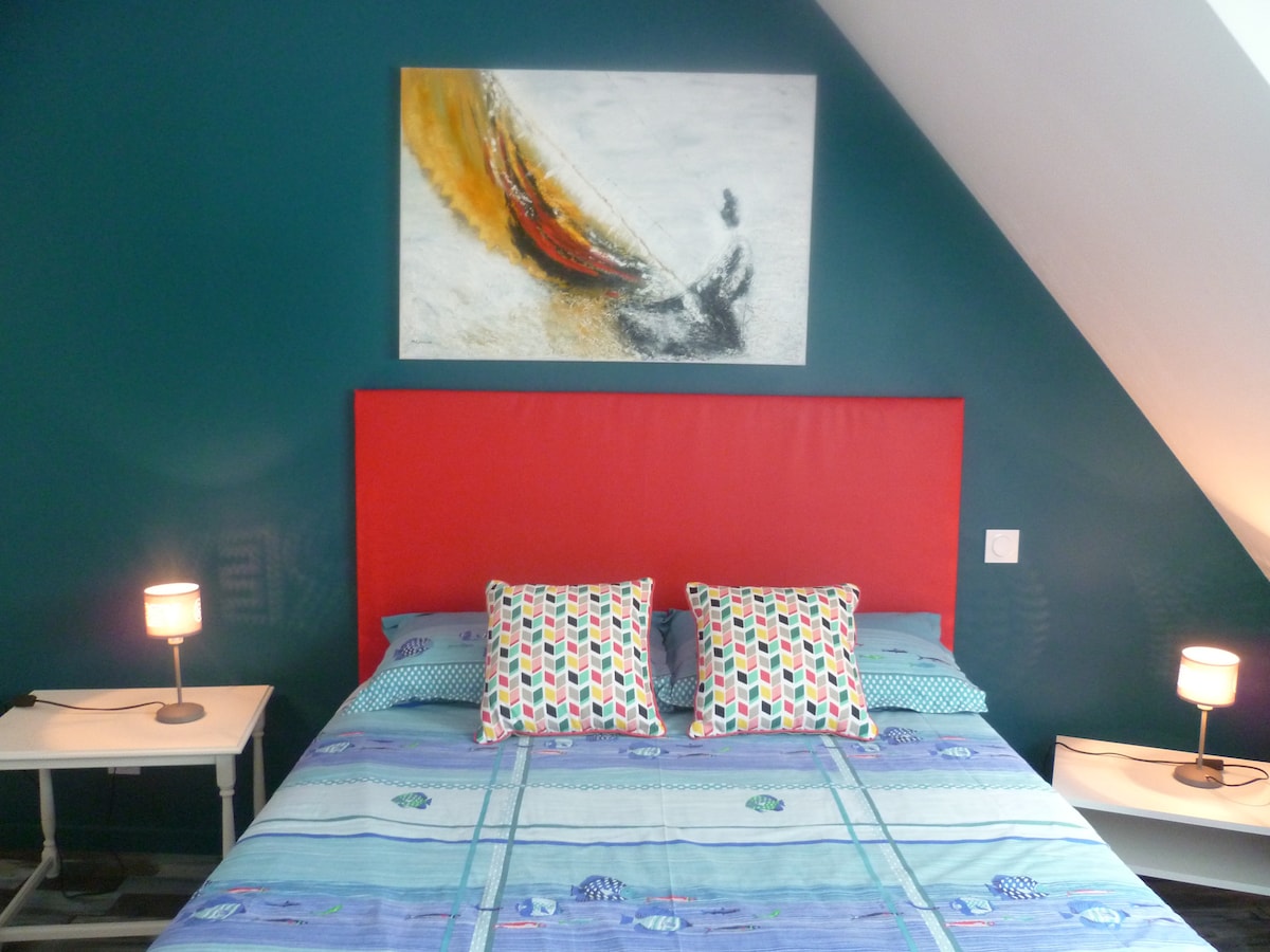 A sleeping area is presented with a spacious bed featuring a vibrant red headboard. Two decorative pillows with playful patterns are arranged against an ocean-themed blanket. Soft lamps are placed on either side of the bed, providing ambient lighting, while a colorful artwork hangs above.