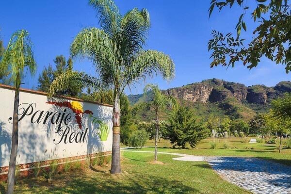 A welcoming entrance is framed by lush greenery and palm trees, leading to a landscaped area. A sign with the word 'Paraje Real' is displayed prominently against a backdrop of rolling hills and clear blue skies.