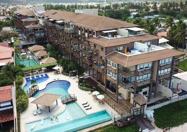 An aerial view of a multi-story complex reveals a vibrant layout featuring an expansive infinity pool and a separate swimming area. Surrounding palm trees provide an oasis-like setting, complemented by thatched gazebos and lounge chairs near the pools, creating inviting spaces for relaxation.
