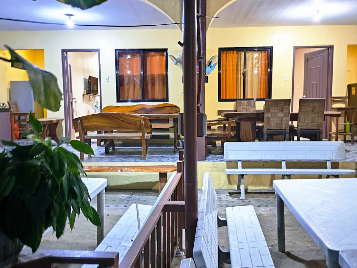The image features a patio area with wooden furniture, including benches and tables. Natural light filters in through large windows, revealing a warm interior. The space is framed by plants, creating a relaxed atmosphere for gatherings.