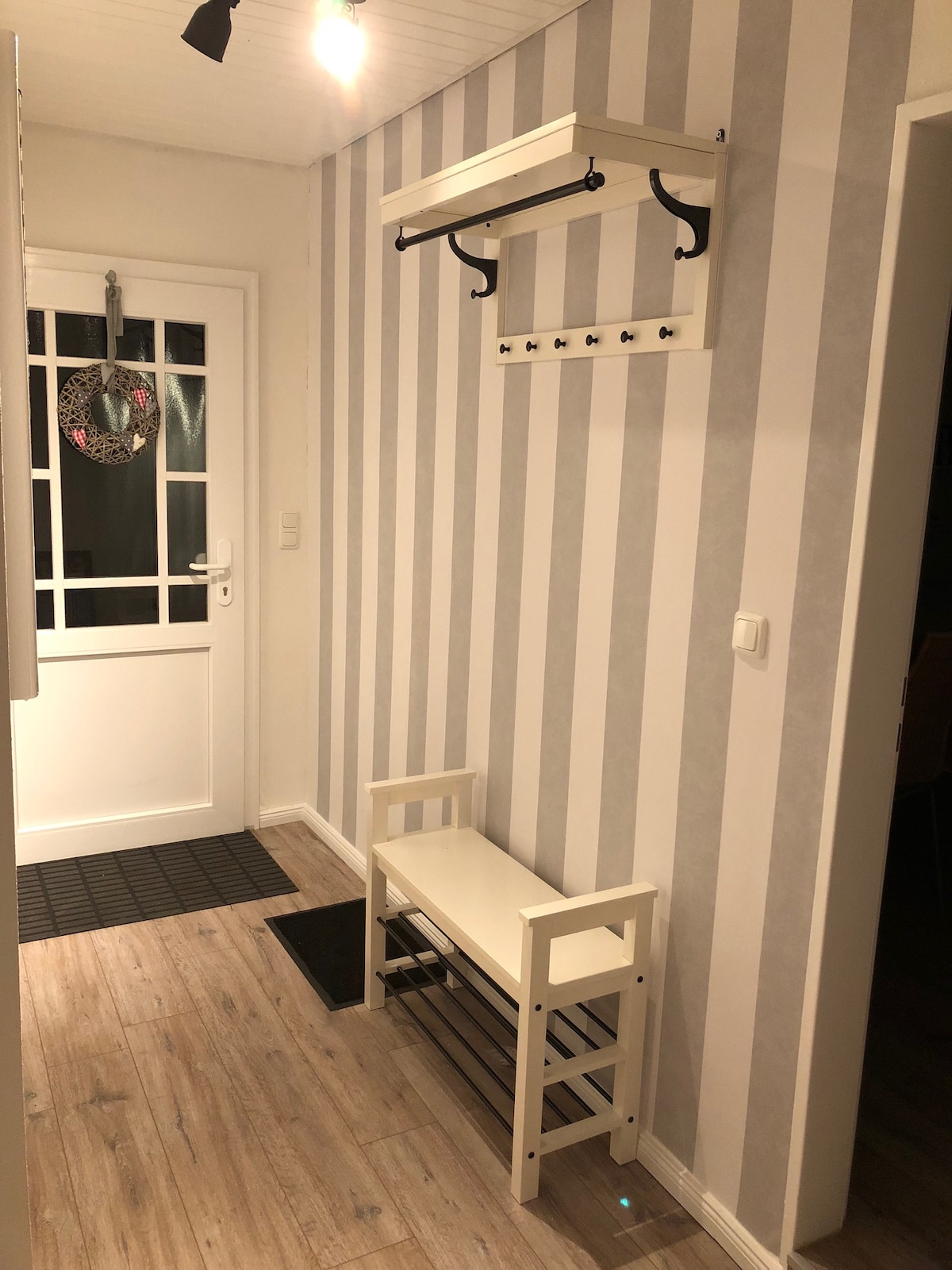 The entryway features light-colored striped walls, a white bench with a lower shelf, and a coat rack with multiple hooks. A doorway leads outside, and there is a mat on the floor, providing a welcoming space for guests upon arrival.