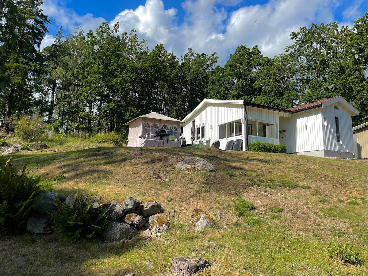 A modern white house is set on a gently sloping lawn, surrounded by lush trees. A large tent with outdoor furnishings is visible nearby. The area features rocky landscaping and greenery, contributing to a serene outdoor setting.