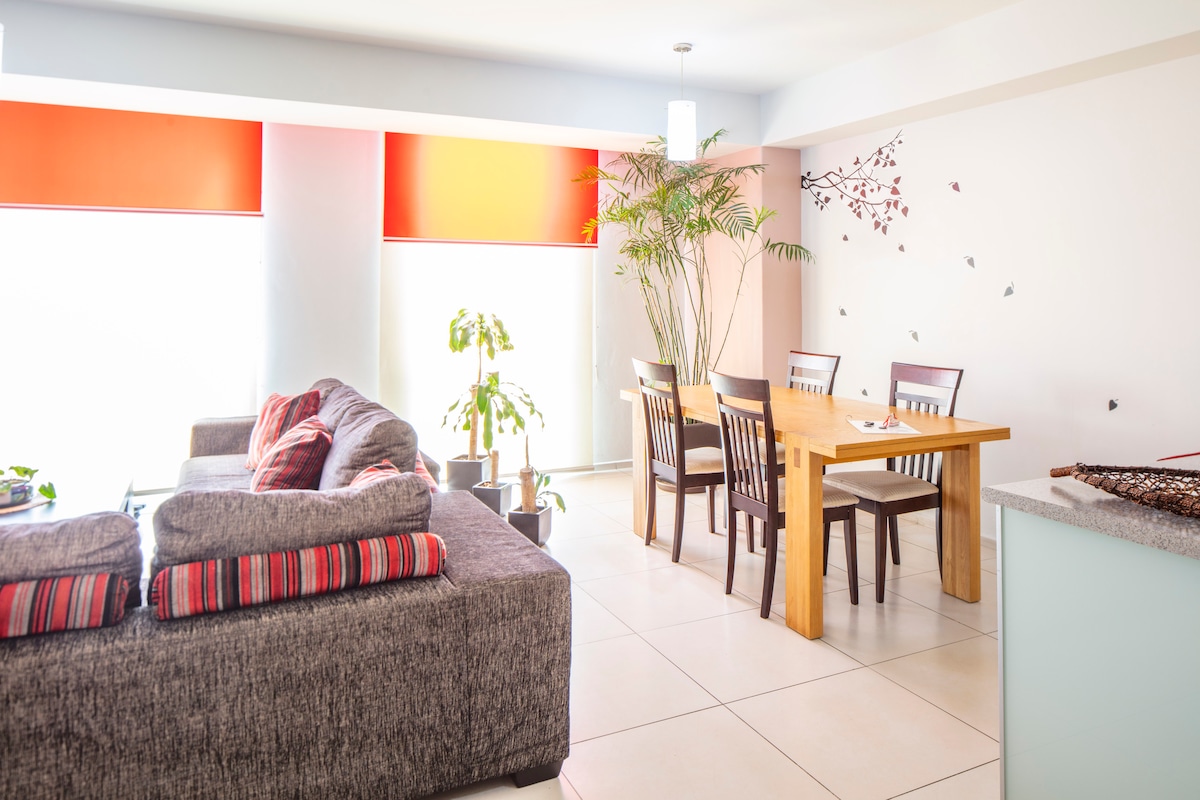 A bright living area features a spacious gray sofa adorned with colorful cushions, alongside a wooden dining table surrounded by four chairs. Large windows with orange shades allow natural light to fill the space, complemented by a few potted plants for a touch of greenery.