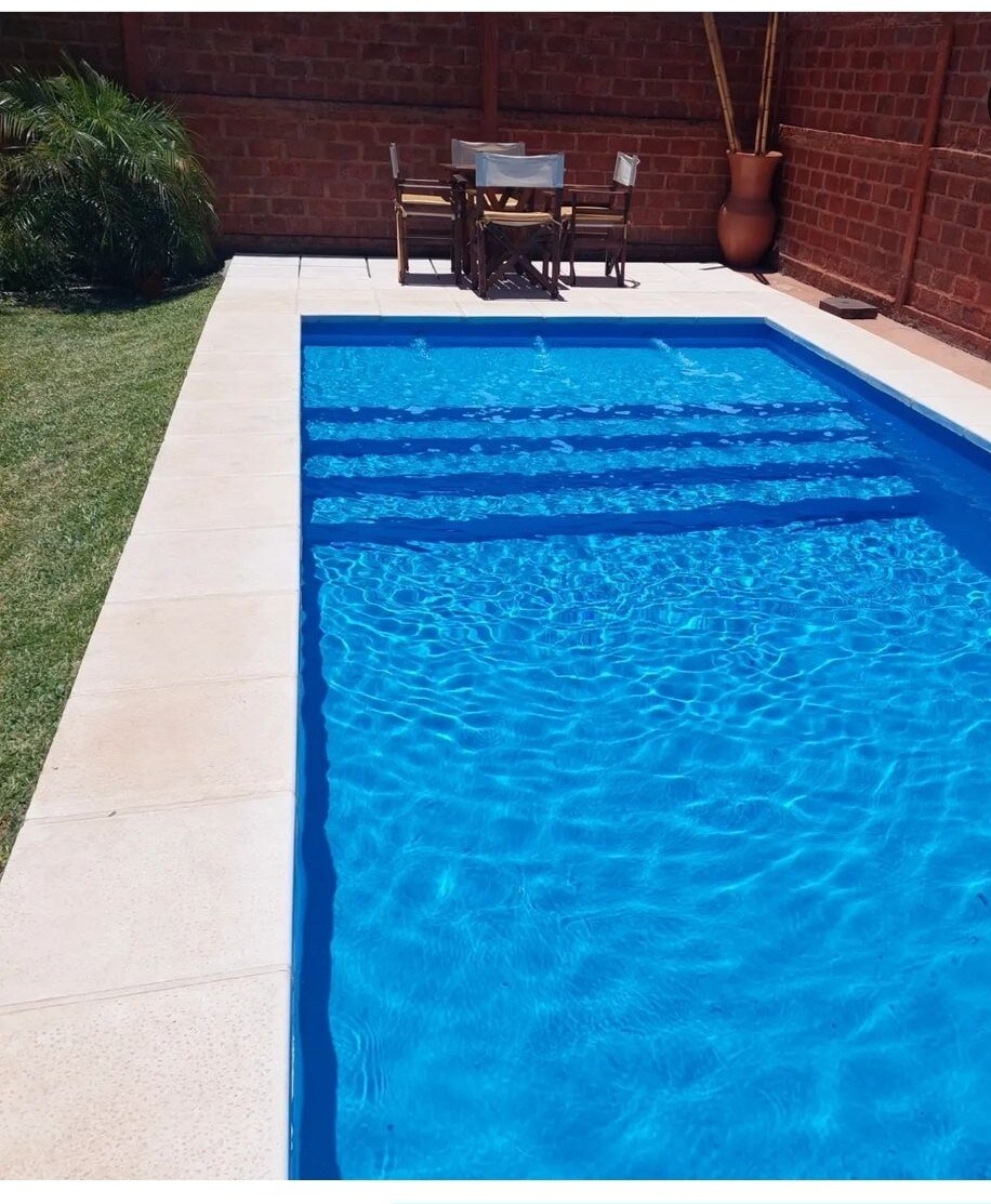 A refreshing swimming pool is surrounded by a clean tiled border, with bright blue water inviting relaxation. A small dining table with four chairs sits nearby, complemented by lush greenery and a decorative pot, providing a serene outdoor space.