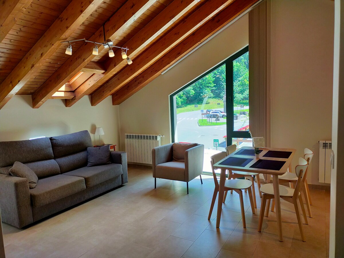 A living area features a cozy gray sofa juxtaposed with a modern dining table surrounded by white chairs. Large windows invite natural light, highlighting the sloped wooden ceiling and showcasing views of the green outdoors.