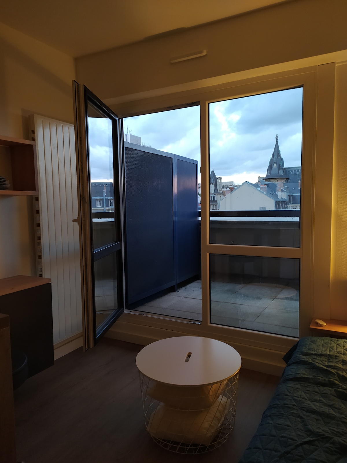 A well-lit studio features a large window and balcony door, providing access to a small outdoor area. The view includes rooftops and a distant steeple. A round table stands beside a cozy bed, enhancing the compact space's functionality and aesthetic.