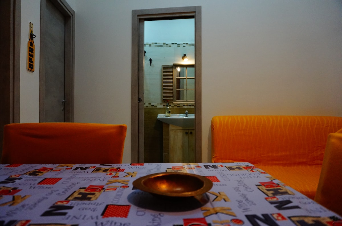 A cozy living area features a colorful table with printed designs, complemented by two bright orange seating areas. A doorway leads to a bathroom illuminated by soft lighting, showcasing modern fixtures and a window with wooden shutters.