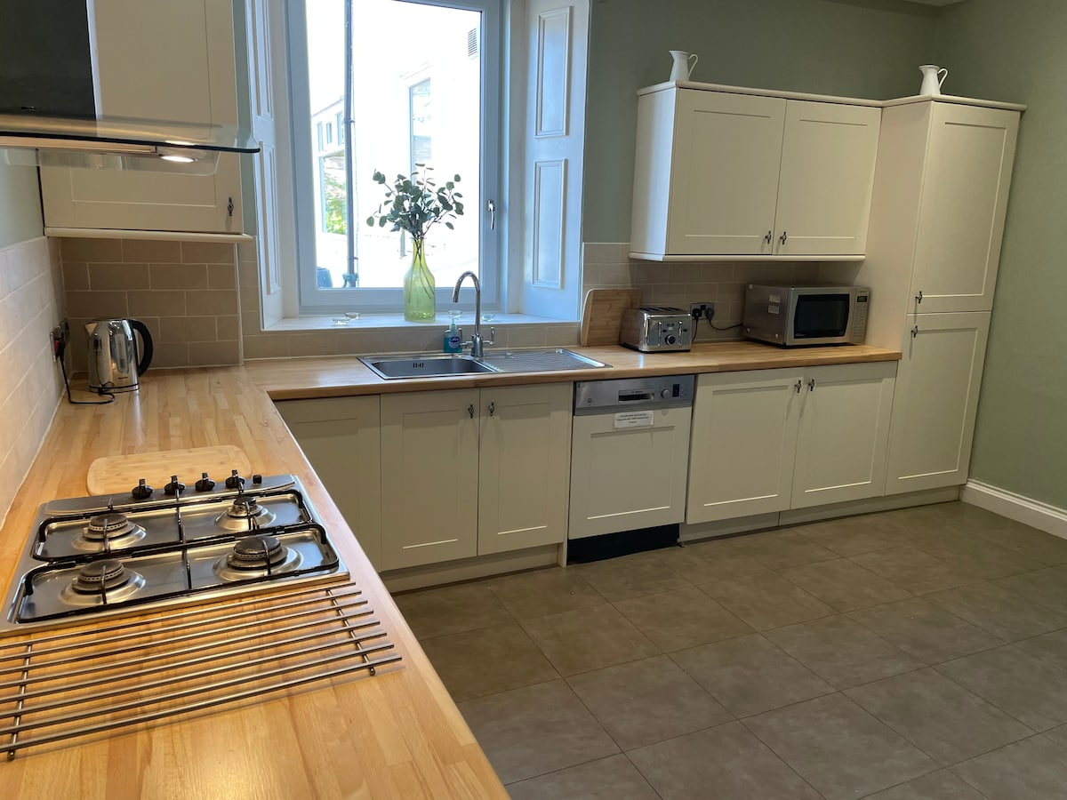 The kitchen features a spacious layout with light-colored cabinetry and a wooden countertop. A gas stove is visible along with a sink and modern appliances, including a microwave and kettle. Natural light enters through a large window, illuminating the functional workspace.