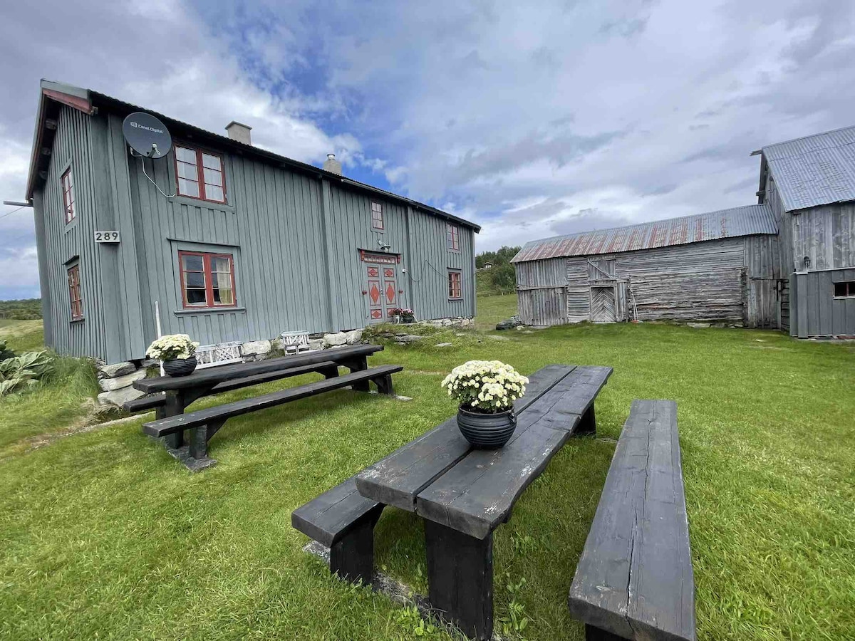 A historic wooden building stands beside a rustic barn, both offering a blend of fading color and durability. Two long picnic tables are set on green grass, each adorned with a planter of white flowers. A cloudy sky looms overhead, enhancing the serene outdoor setting.
