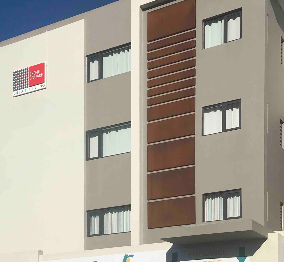 A multi-story building is shown, featuring a modern façade with a mix of neutral colors. Large windows adorned with white curtains allow natural light to enter. The building's entrance is partially visible, indicating access to the interior.