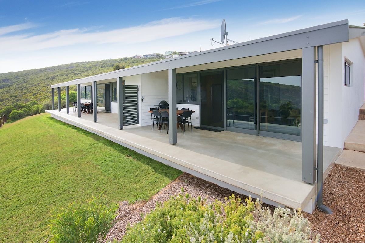 The exterior of the apartment is shown, featuring a wide, covered patio that provides panoramic views of the surrounding hills. Sleek glass doors open to the interior, while a spacious outdoor dining area is visible. Lush greenery complements the modern architecture.