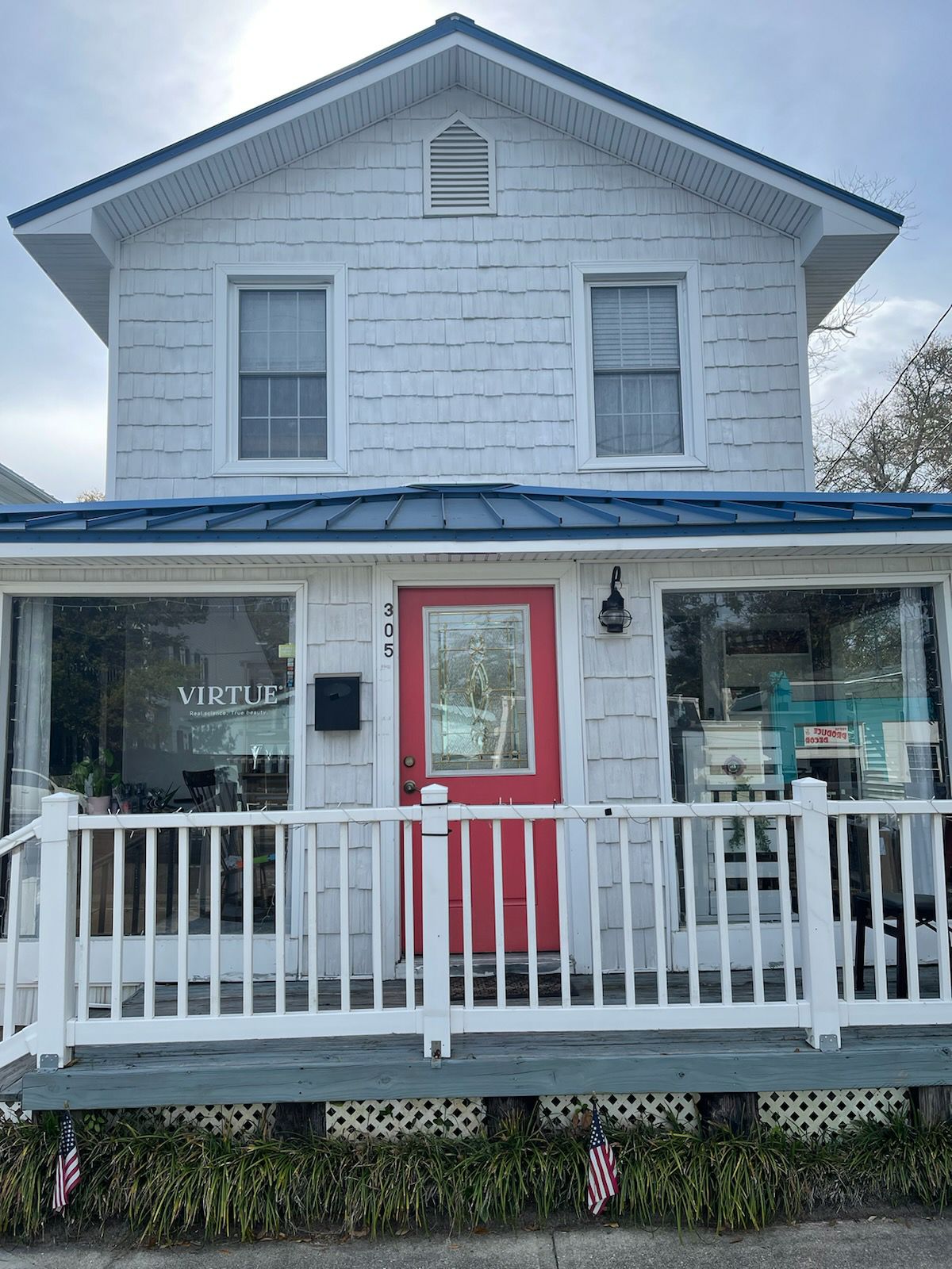 A charming two-story home with white shingle siding and a bright red front door is shown. The welcoming porch features a railing and small flags positioned along the base, while large windows reveal the interior space and inviting atmosphere.