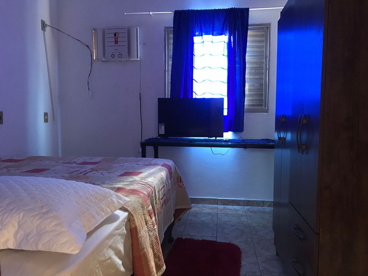 A bright sleeping area features a spacious bed covered with a patterned quilt. A dark wood wardrobe stands nearby, and a small desk with a television is positioned against the wall. Natural light enters through a window dressed in blue curtains.