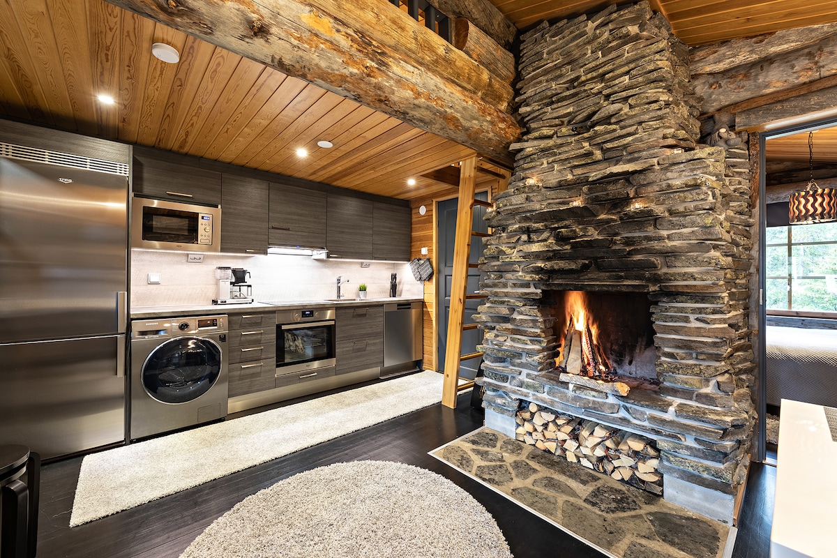 A modern kitchen is set against a rustic stone fireplace, featuring stainless steel appliances and contemporary cabinetry. A soft rug lies on the dark floor, while natural light streams in from nearby windows, illuminating the warm wooden beams overhead.