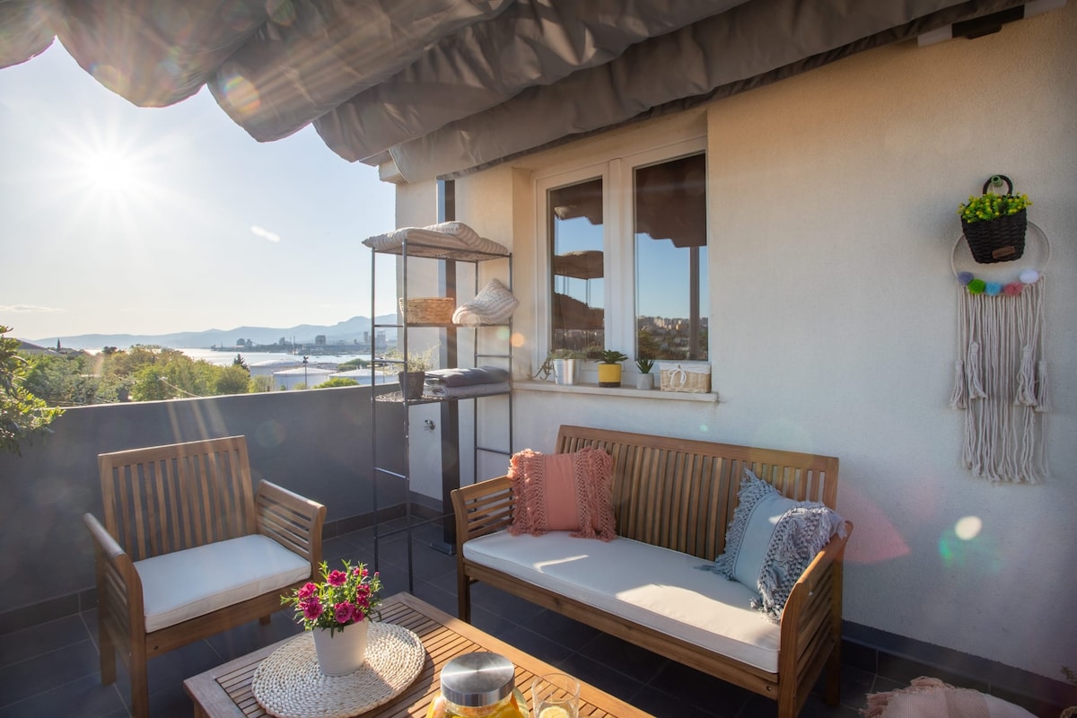 A relaxing outdoor terrace is featured with wooden seating arrangements and a table, surrounded by greenery. Sunlight creates a warm atmosphere, while the backdrop includes a panoramic view of the sea and distant islands. Umbrellas provide shade, ensuring comfort throughout the day.