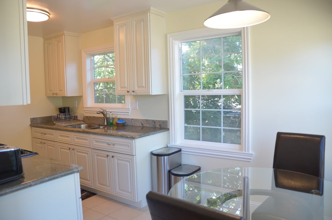 A bright kitchen area features white cabinetry and granite countertops. Natural light enters through dual windows overlooking greenery. A round glass dining table is positioned beside the windows, paired with dark, upholstered chairs. Modern appliances and a stylish light fixture complete the space.