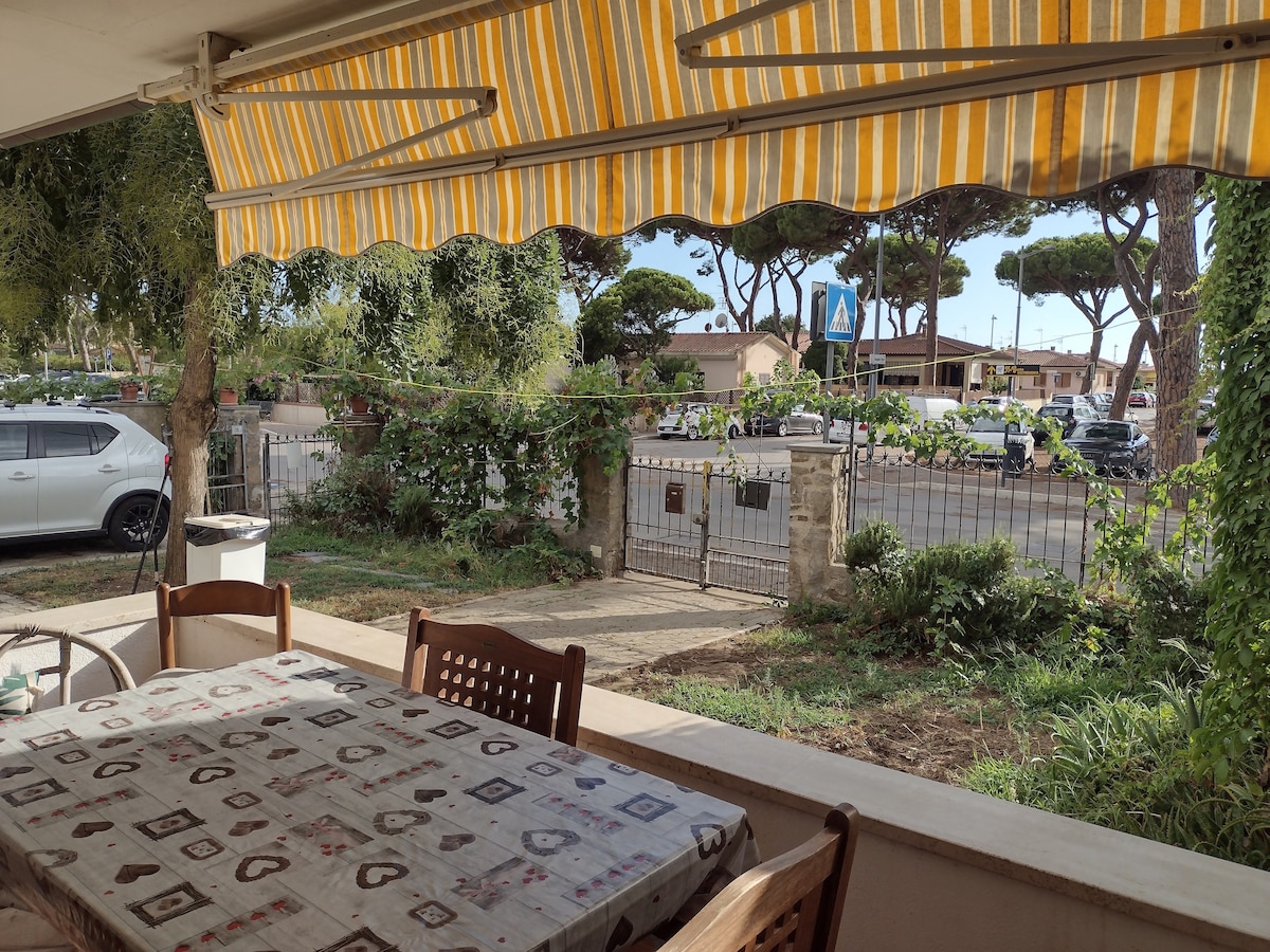 A covered outdoor dining area is presented with a patterned tablecloth and wooden chairs. The view includes a landscaped garden and a gated entryway, framed by tall trees. A parked car is visible nearby, along with other vehicles along a nearby street.