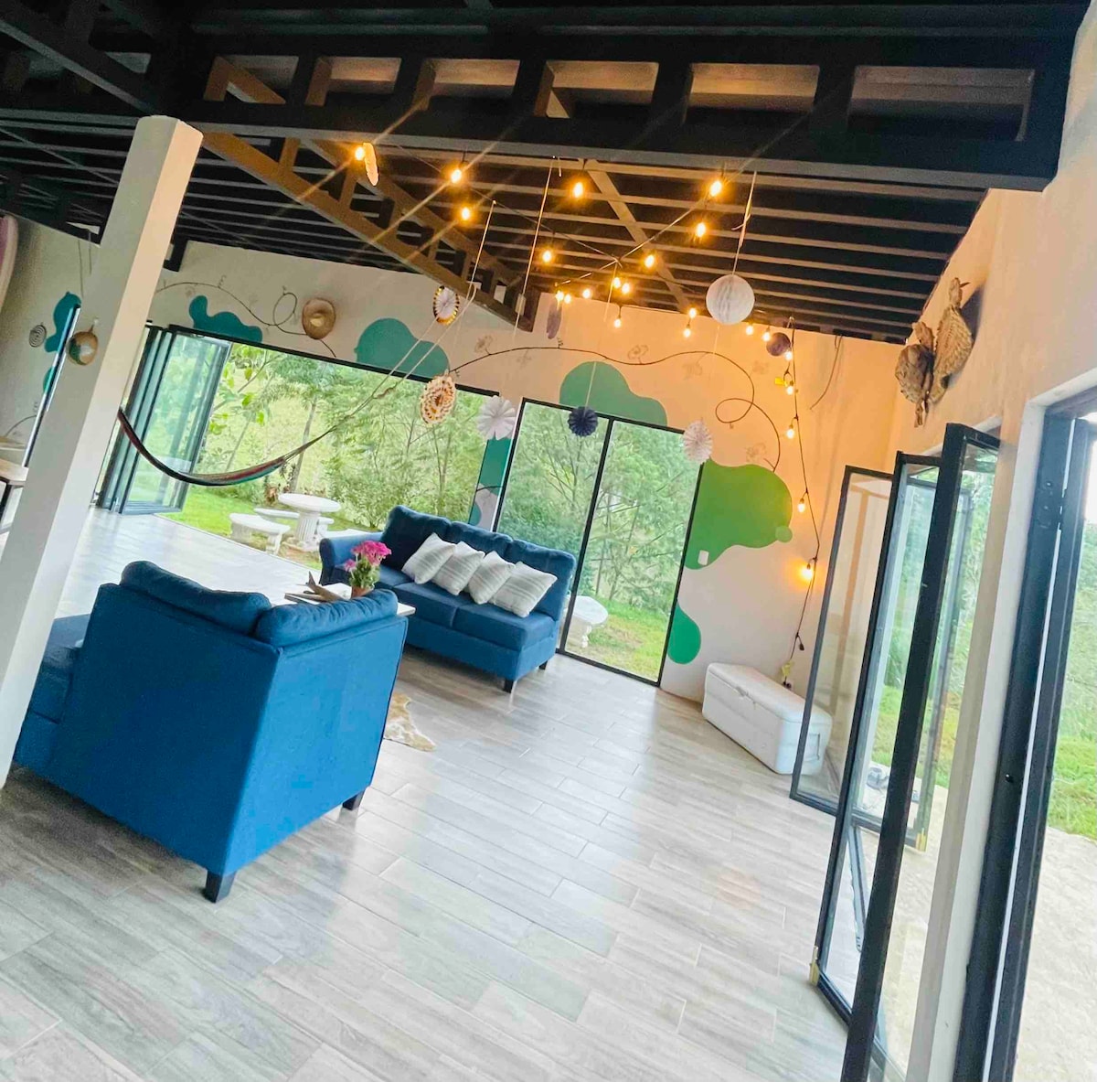 A spacious living area is illuminated by warm string lights, featuring large glass doors that connect the indoors to the natural surroundings. A blue sofa with decorative pillows is positioned thoughtfully, complemented by a hammock and colorful wall art enhancing the vibrant atmosphere.