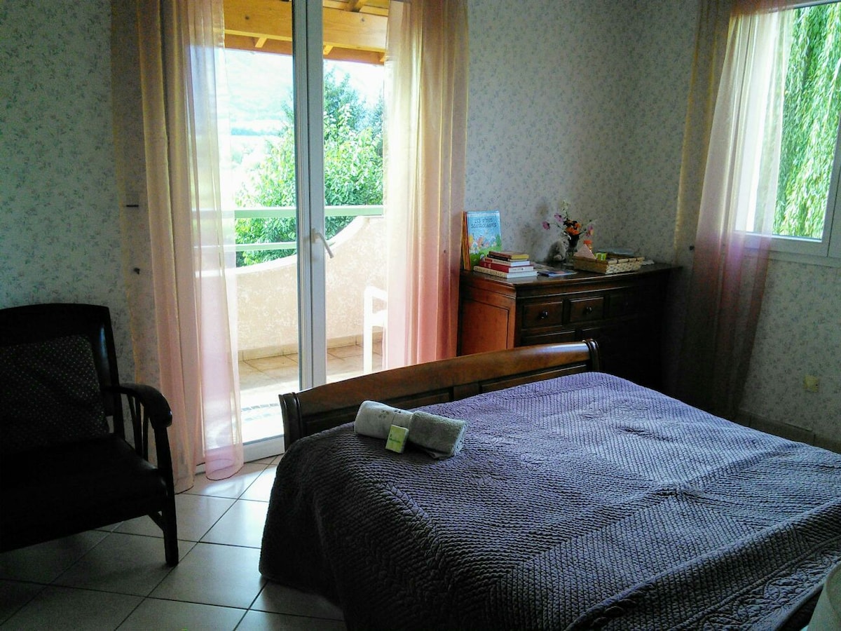 A bright bedroom is presented with a spacious bed covered in a textured blanket. Natural light pours in from glass doors leading to a balcony, framed by sheer curtains. A wooden dresser and a chair are positioned in the room, contributing to a tranquil atmosphere.
