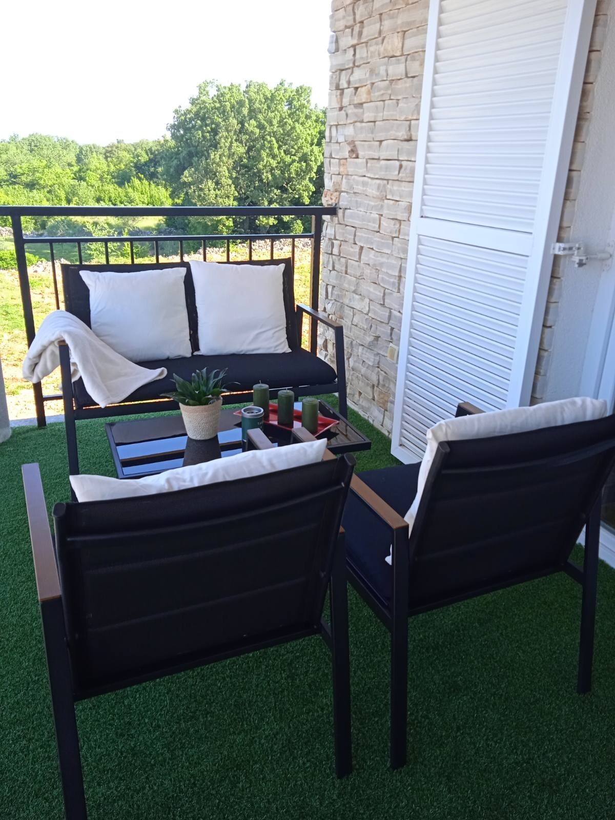 A balcony space features two black chairs adjacent to a compact table, inviting relaxation. A potted plant and a tray with candles add a touch of charm. The balcony is surrounded by greenery, providing a serene outdoor setting.