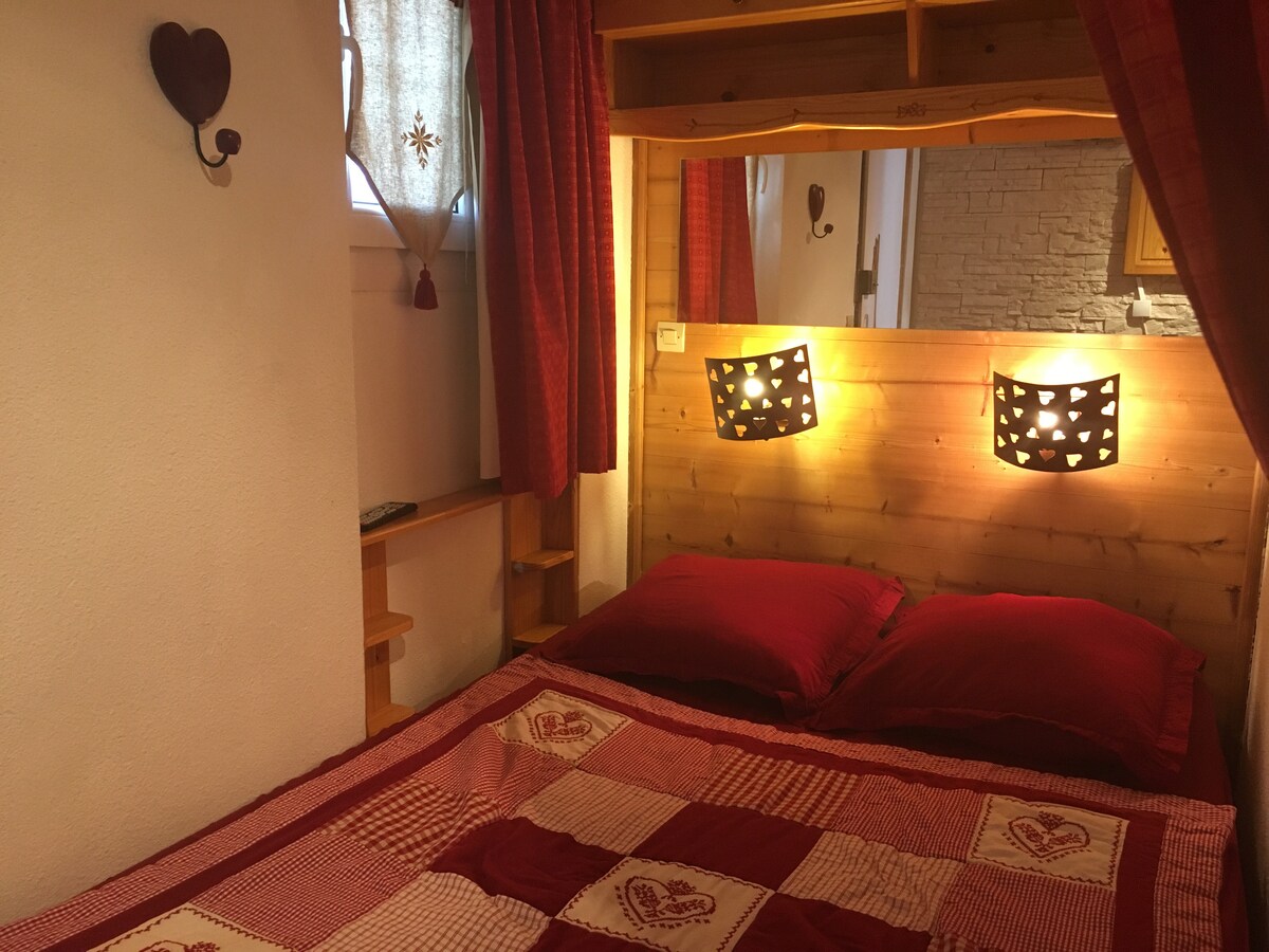 A cozy sleeping area is presented, featuring a rustic wood headboard with decorative lighting. The bed is dressed in red and white patterned linens, while sheer curtains allow soft light to filter in. Shelving is positioned beside the bed, enhancing functionality.