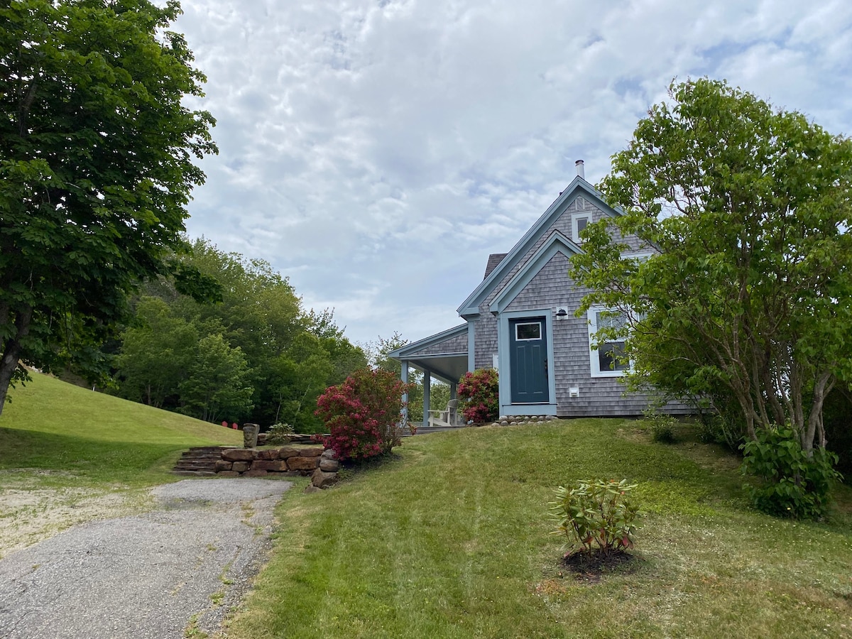 A charming historic home is nestled among greenery, showing a well-maintained lawn with a gravel driveway. The exterior features grey shingles and decorative trim. Flowering shrubs add color to the landscape, while a gentle slope leads to the surrounding wooded area.