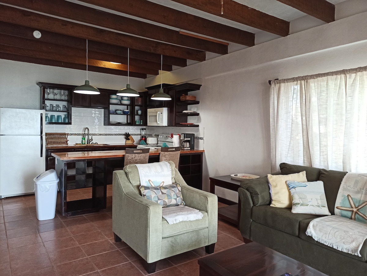 The living space features exposed wooden beams and a fully equipped kitchen with ample storage. A comfortable green armchair and a dark brown coffee table complement the beige sofa, which is adorned with decorative pillows. Natural light flows in through sheer curtains, enhancing the inviting ambiance.