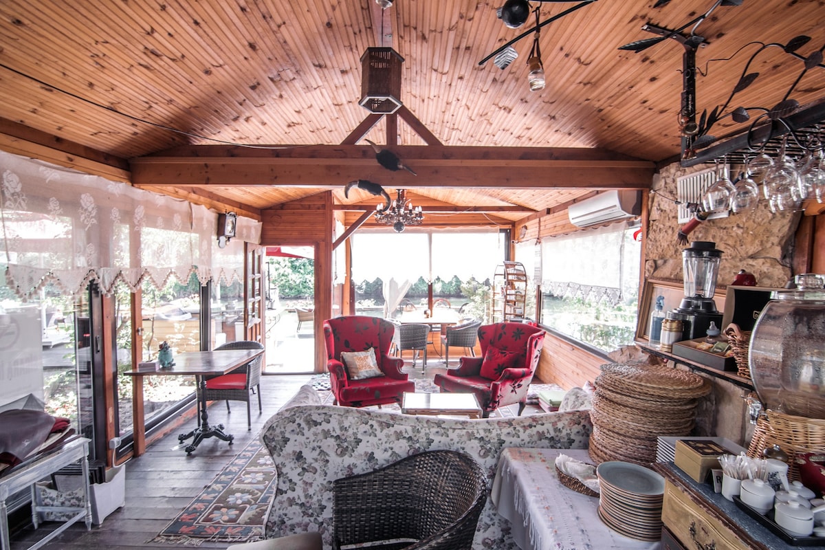 A cozy interior space features wooden walls and ceiling, complemented by large windows allowing natural light. Soft seating is arranged with red armchairs and a patterned rug. Decorative items, including woven baskets and a chandelier, enhance the inviting atmosphere of the room.