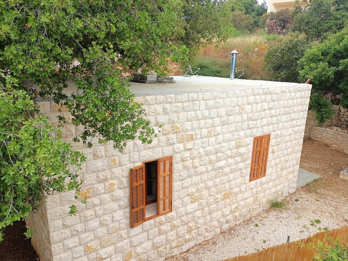 The exterior of a stone house is presented, featuring wooden shutters on the windows and a flat roof partially shaded by overhanging trees. A natural garden surrounds the structure, enhancing the rustic appeal.