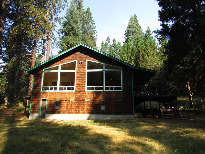 A spacious wooden home is set among tall pine trees, featuring a large deck and expansive windows that allow natural light to fill the interior. The building is designed with a sloping roof and stands on a grassy area, surrounded by nature.