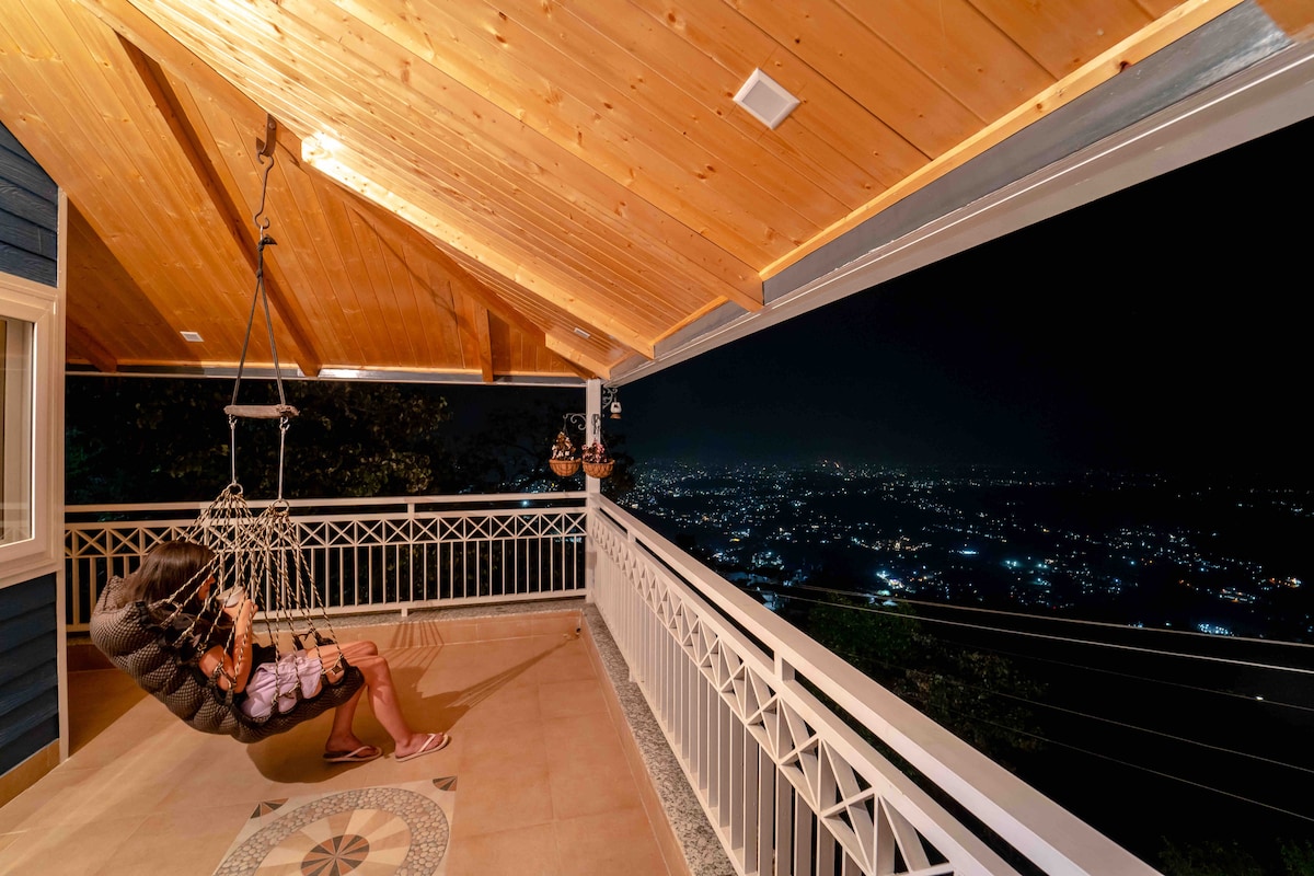 A spacious balcony features a woven hanging chair, providing a comfortable seating option. The ceiling beams are made of wood, and the area is illuminated, showcasing a panoramic view of the twinkling city lights of Dehradun at night.