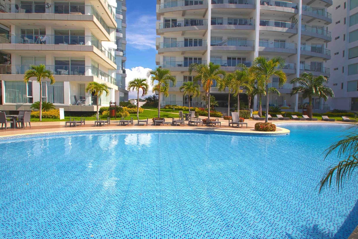 A large swimming pool with clear blue water is surrounded by well-maintained greenery and lounge chairs. The architectural design of adjacent buildings features multiple stories and landscaped areas, providing a bright and inviting space for relaxation.