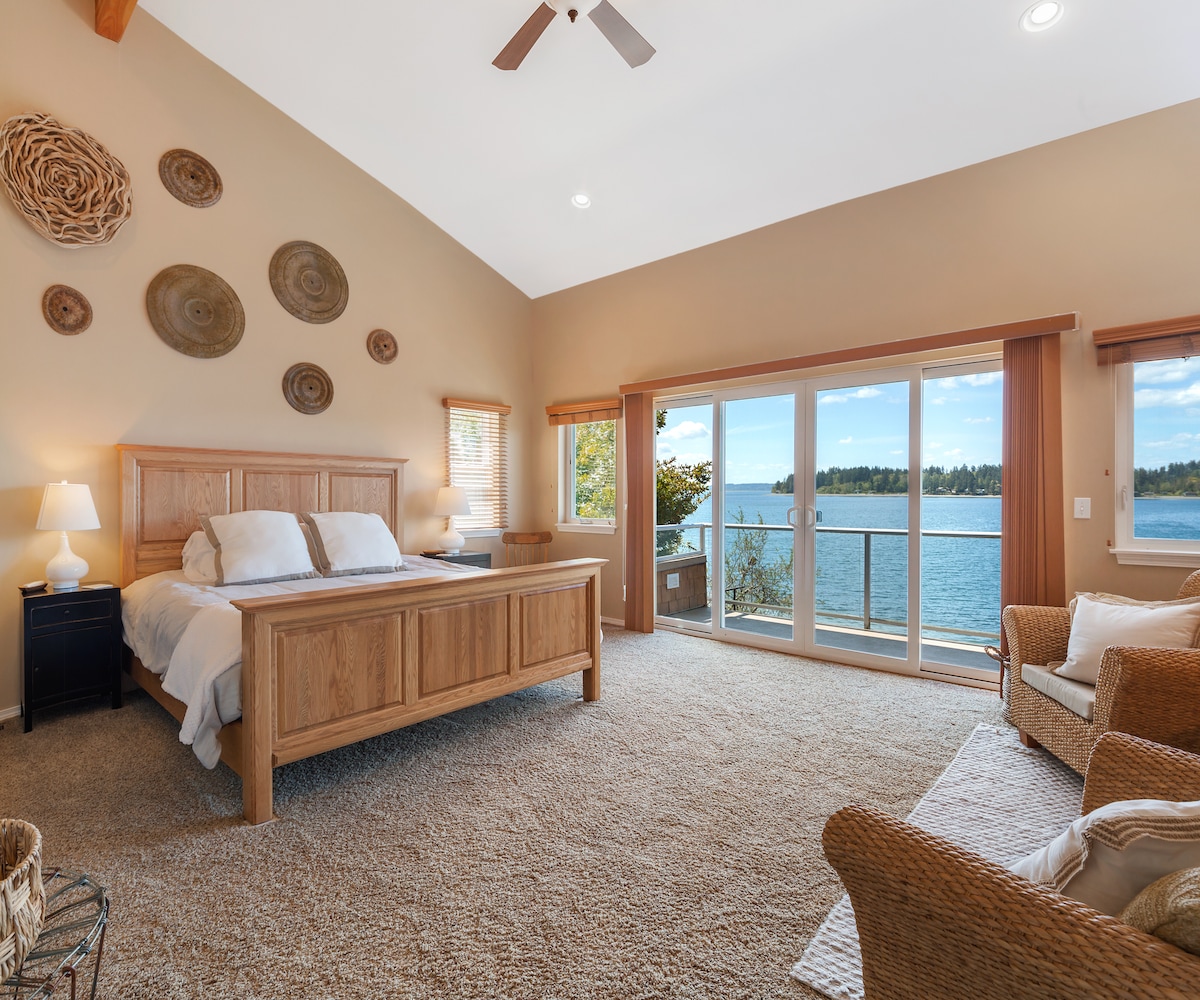 A spacious master bedroom features a large wooden bed facing sliding glass doors that open to a private deck with water views. Decorative wall accents and natural light create a serene environment. Comfortable seating options are positioned near the window for relaxation.
