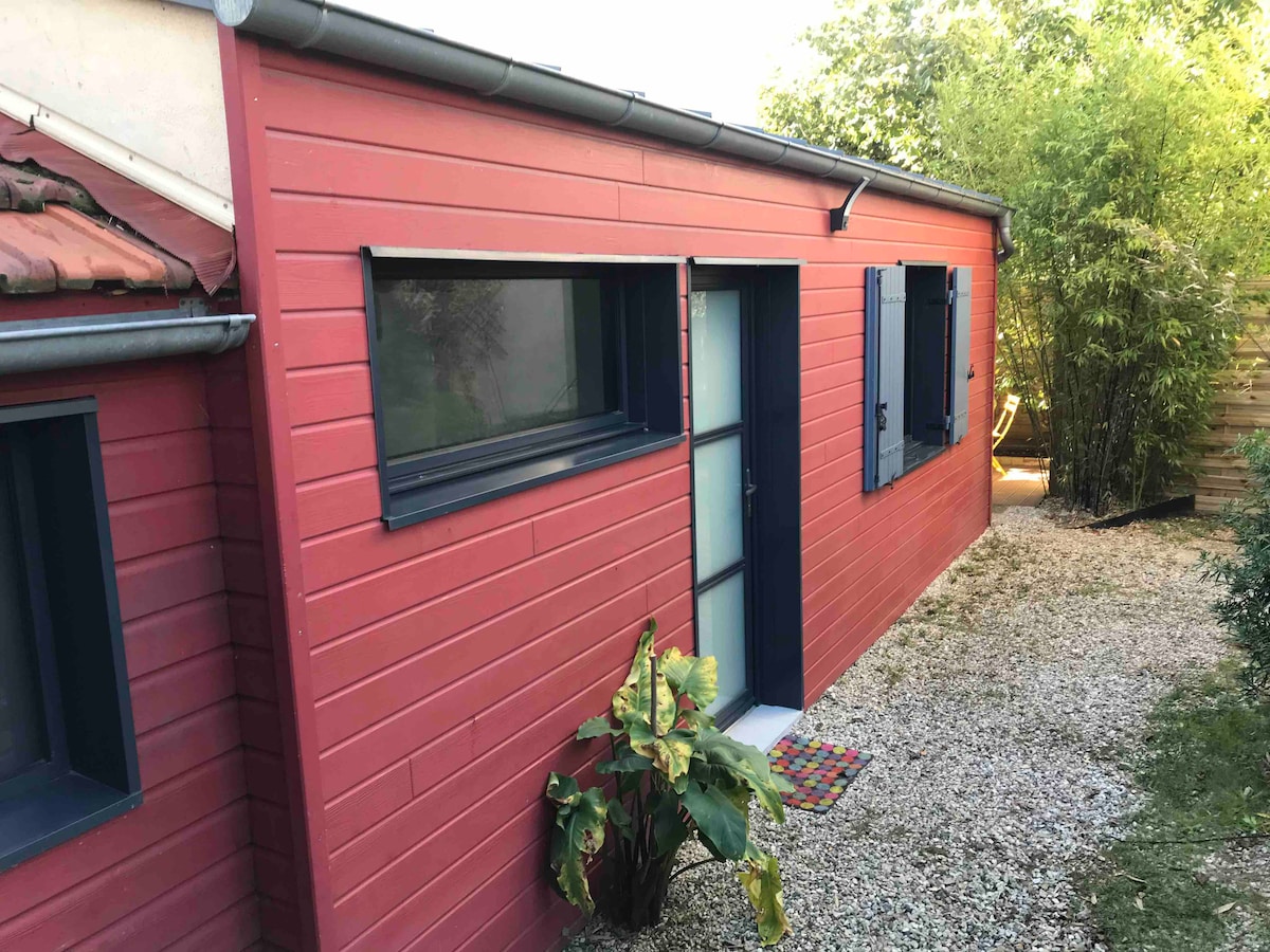 A modern red exterior of the studio is highlighted by dark blue window frames and a sleek door. A vibrant mat welcomes visitors at the entrance, and a path lined with greenery leads to the door, enhancing the natural surroundings.