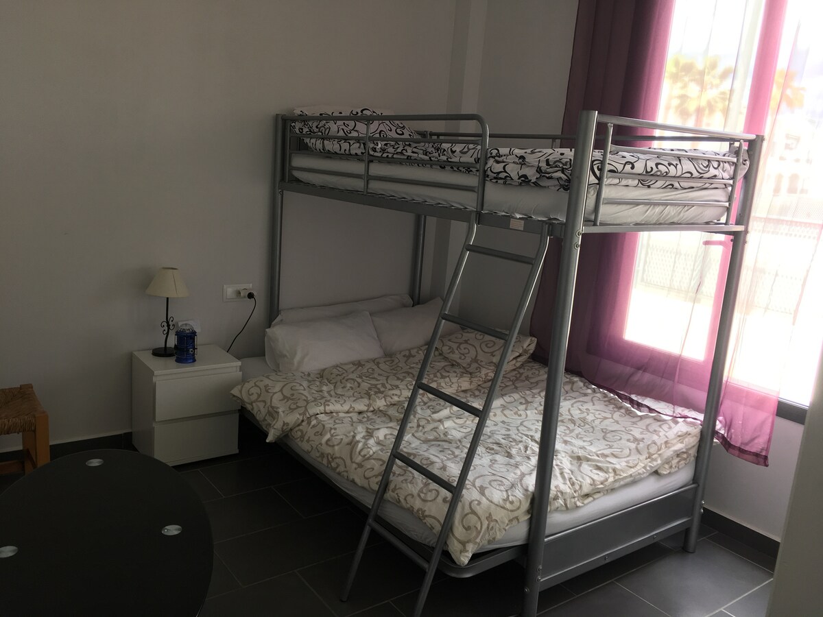 A compact bedroom showcases a bunk bed with a top mattress and a spacious bottom bed. A nightstand is positioned beside the lower bed, illuminated by a small lamp. Light filters in through large windows adorned with sheer curtains, enhancing the room's brightness.
