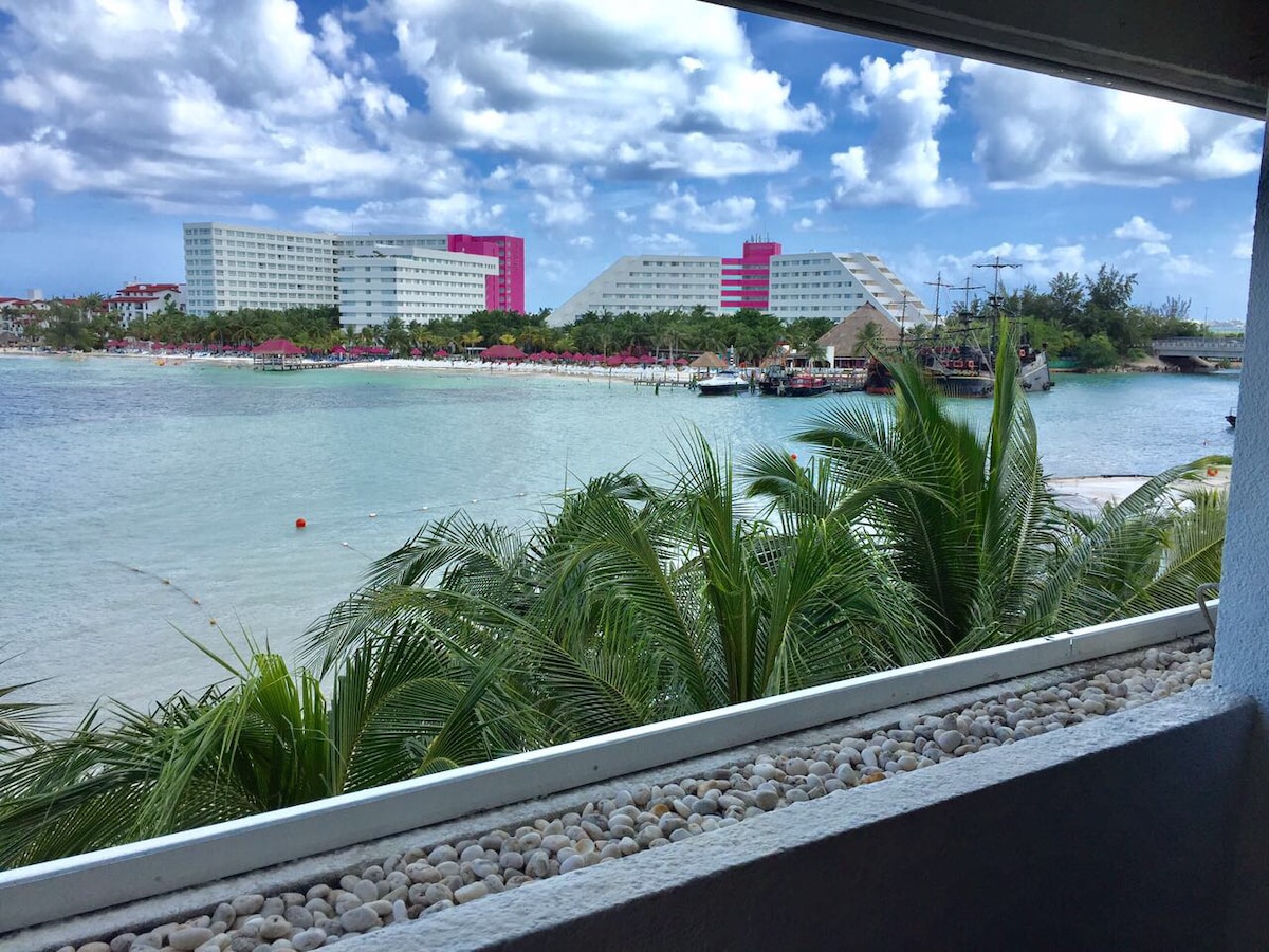 A view showcases the turquoise bay framed by palm leaves. In the background, modern buildings are visible, with a vibrant pink structure and terraced architecture blending into the coastal scenery. The inviting water reflects the bright sky dotted with clouds.