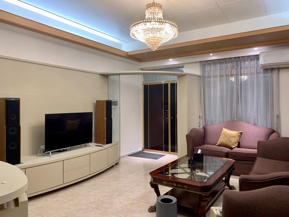 A living area features a television on a light-colored cabinet, accompanied by two speakers. A coffee table sits at the center, surrounded by a sofa and an armchair. Natural light filters through sheer curtains, and a chandelier hangs from the ceiling, illuminating the space.